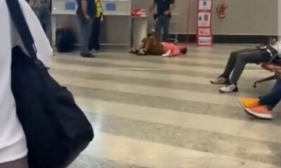 Mulher deitada no chão de um aeroporto, cercada por passageiros e funcionários, em frente ao portão B30 no terminal de embarque