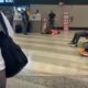 Mulher deitada no chão de um aeroporto, cercada por passageiros e funcionários, em frente ao portão B30 no terminal de embarque
