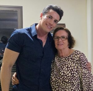 Homem de camisa azul abraça uma mulher de óculos e vestido estampado; ambos sorriem para a câmera em um ambiente interno