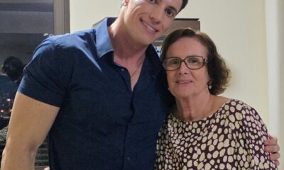 Homem de camisa azul abraça uma mulher de óculos e vestido estampado; ambos sorriem para a câmera em um ambiente interno