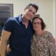 Homem de camisa azul abraça uma mulher de óculos e vestido estampado; ambos sorriem para a câmera em um ambiente interno