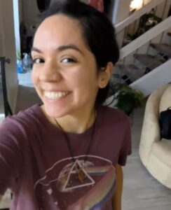 Mulher jovem sorrindo em ambiente interno, usando camiseta roxa estampada e colar, antes de sofrer acidente em trilha no Arizona