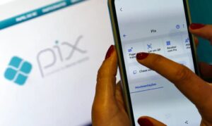 Pessoa utilizando o aplicativo de Pix no celular, com tela exibindo opções de pagamento; ao fundo, o logotipo do sistema Pix em uma tela.