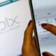 Pessoa utilizando o aplicativo de Pix no celular, com tela exibindo opções de pagamento; ao fundo, o logotipo do sistema Pix em uma tela.