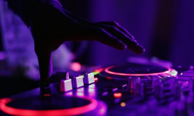 Mão de DJ manipulando mesa de som iluminada com luzes roxas e vermelhas durante evento noturno.