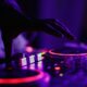 Mão de DJ manipulando mesa de som iluminada com luzes roxas e vermelhas durante evento noturno.