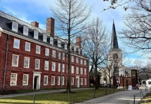 Prédio histórico de tijolos vermelhos e torre de igreja ao fundo no campus da Universidade Harvard, com árvores sem folhas e céu azul claro.