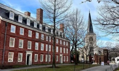 Prédio histórico de tijolos vermelhos e torre de igreja ao fundo no campus da Universidade Harvard, com árvores sem folhas e céu azul claro.