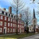 Prédio histórico de tijolos vermelhos e torre de igreja ao fundo no campus da Universidade Harvard, com árvores sem folhas e céu azul claro.