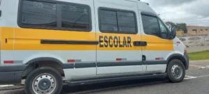 Imagem de uma van escolar branca com faixa amarela estacionada na rua; veículo semelhante ao usado para transporte de crianças.