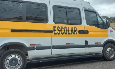 Imagem de uma van escolar branca com faixa amarela estacionada na rua; veículo semelhante ao usado para transporte de crianças.