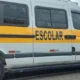 Imagem de uma van escolar branca com faixa amarela estacionada na rua; veículo semelhante ao usado para transporte de crianças.