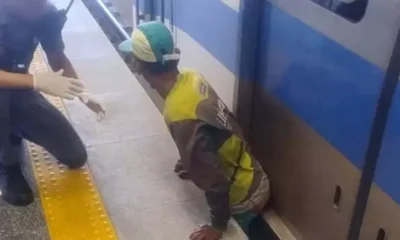 Homem com uniforme de agente de limpeza está preso entre o trem e a plataforma do metrô, enquanto policial militar com luvas o auxilia durante resgate. Cena ocorreu na estação Campinas de Pirajá, em Salvador