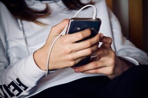 Pessoa com blusa branca segura um celular com fone de ouvido conectado, usando o aparelho com as duas mãos