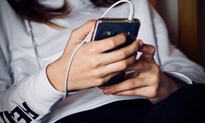 Pessoa com blusa branca segura um celular com fone de ouvido conectado, usando o aparelho com as duas mãos