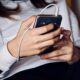 Pessoa com blusa branca segura um celular com fone de ouvido conectado, usando o aparelho com as duas mãos