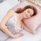 Mulher deitada em uma cama com roupa de dormir de cetim, descansando com expressão tranquila sobre um travesseiro rosa claro.