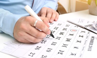 Pessoa com camisa azul riscando dias em um calendário de mesa com caneta preta, representando o acompanhamento de datas e feriados.