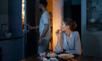 Mulher sentada à mesa comendo enquanto homem abre a geladeira em um ambiente de cozinha com pouca luz; há pratos com lanches e sobremesas sobre a mesa.