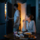Mulher sentada à mesa comendo enquanto homem abre a geladeira em um ambiente de cozinha com pouca luz; há pratos com lanches e sobremesas sobre a mesa.