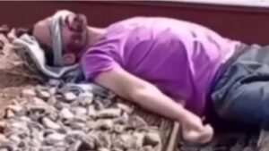 Homem de camiseta roxa e calça escura deitado entre trilhos de trem, com a cabeça apoiada em panos e os olhos cobertos, em cena capturada de vídeo feito em estação da CPTM.