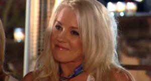 Tamika Chesser sorrindo em cena do reality show As Gostosas e os Geeks, com cabelo loiro solto e maquiagem leve.