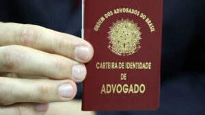 Mão segurando uma carteira vermelha da Ordem dos Advogados do Brasil (OAB), com o brasão nacional e a inscrição "Carteira de Identidade de Advogado" em letras douradas.