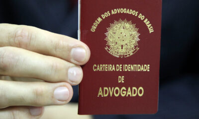 Mão segurando uma carteira vermelha da Ordem dos Advogados do Brasil (OAB), com o brasão nacional e a inscrição "Carteira de Identidade de Advogado" em letras douradas.