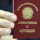 Mão segurando uma carteira vermelha da Ordem dos Advogados do Brasil (OAB), com o brasão nacional e a inscrição "Carteira de Identidade de Advogado" em letras douradas.