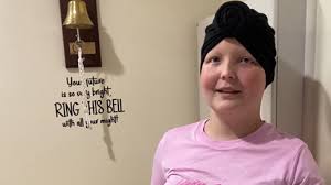 Garota sorridente usando turbante preto toca o sino da cura no hospital, com frase motivacional na parede ao lado, após vencer o câncer.