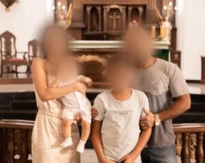 Foto de uma família composta por pai, mãe e dois filhos pequenos, com os rostos desfocados, posando juntos em frente a um altar de igreja.
