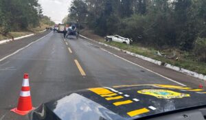 Foto de uma rodovia após um acidente de trânsito, mostrando um carro branco destruído fora da pista.