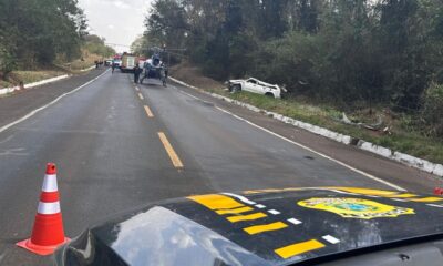 Foto de uma rodovia após um acidente de trânsito, mostrando um carro branco destruído fora da pista.