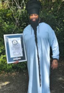 Homem negro com vestimenta azul e com a barba mais longa do mundo segurando o certificado do Guinness World Records.