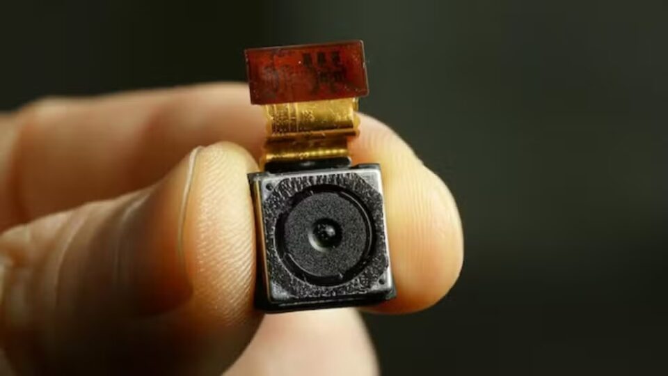 Imagem mostra uma mão segurando com o indicador e o polegar uma câmera pequena.
