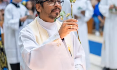 Padre de vestimentas brancas segurando uma rosa com a mão direita.